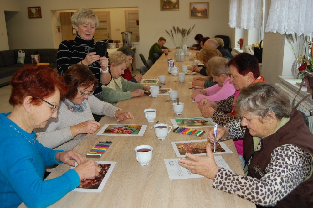 Uczestnicy zajęć siedzą przy stole i malują kolorowe prace.