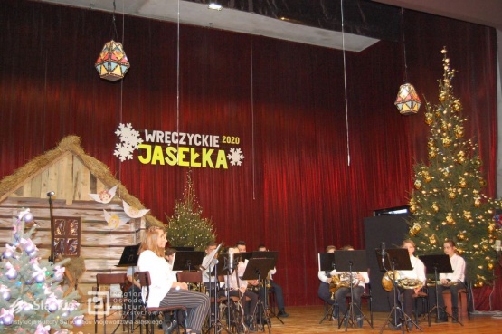 Scena Jasełek, widoczna choinka, napis Wręczyckie Jasełka 2020, zawieszony na czerwonej kotarze, stajenka drewniana oraz muzycy siedzący na krzesłach trzymający instrumenty muzyczne.