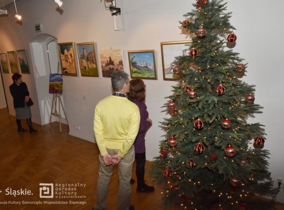 Trójka ludzi oglądających wystawę fotografii powieszonych na ścianach. W prawym rogu widoczna stojąca choinka ubrana w czerwono-złote bombki i złote światełka.