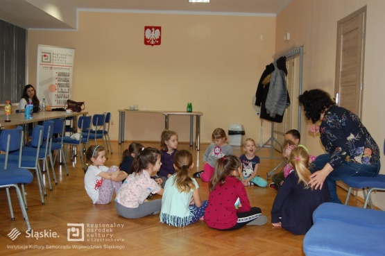 Dzieci siedzą na podłodze i przysłuchują się wskazówkom udzielanym przez prowadzącą warsztaty teatralne.