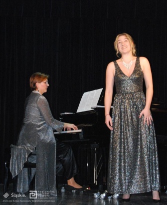  Kobieta w srebrnej sukni z kryształowym naszyjnikiem stoi przy pianinie i śpiewa. Obok inna kobieta gra na pianinie