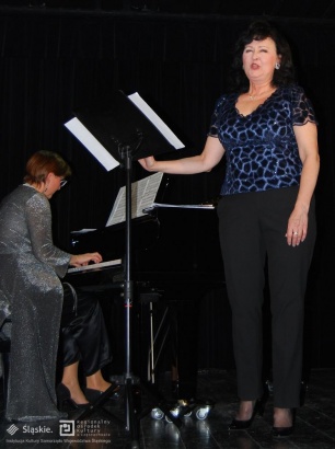 Kobieta na scenie stoi przed statywem z kartką i śpiewa. Obok niej pianistka.