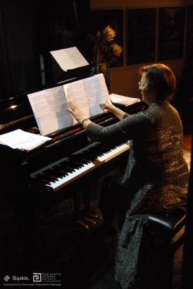 Pianino wraz z pianistką, która zmienia nuty. Pianistka ubrana jest w srebrną świecącą suknię. Za pianiną widnieją kwiaty a za nimi ściana, na której wiszą obrazy.