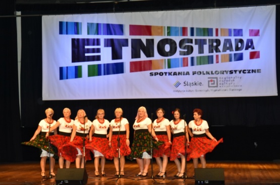 Kobiety tańczące i śpiewające na scenie ubrane są w stroje ludowe. W tle na ekranie projektora widnieje napis Etnostrada Spotkania Folklorystyczne.