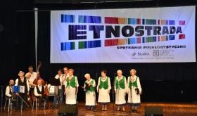 Kobiety w biało-zielonych strojach ludowych śpiewają na scenie. W tle orkiestra składająca się z muzyków.