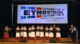 Kobiety w biało-zielonych sukniach śpiewają na scenie, obok nich stoi kobieta grająca na skrzypcach i mężczyzna grający na akordeonie.