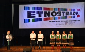 Cztery kobiety ubrane na zielone, dwóch mężczyzn ubranych na biało dają pokaz śpiewu ludowego. Obok mężczyzna, który przygrywa na akordeonie.