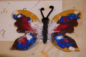 Motyl stworzony z drwna i pomalowany farbami do drewna na kolory: zółty, czerwony, niebieski, biały i czarny.