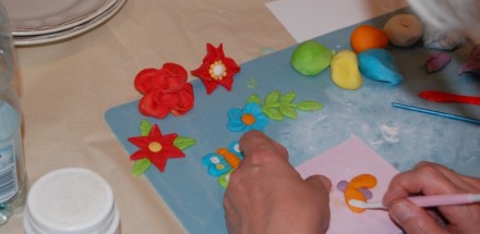 Praca plastyczna uczestnika warsztatów - kolorowe kwiaty wykonane z modeliny naklejone na niebieską kartkę.