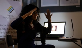 Jolanta Rycerska w trakcie wykładu z fotografii. Lewa ręka jest zgięta w łokciu i lekko uniesiona do góry.