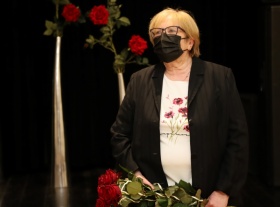 Kobieta trzymająca czerwone róże w rękach. Jest ubrana w czarny żakiet i maseczkę ochronną. W tle widać  czerwone róże w wazonach.