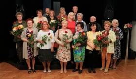 Zdjęcie grupowe eleganckich kobiet ustawionych przed sceną. Wszystkie mają w rękach bukiety kwiatów.