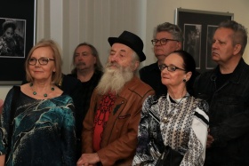 Artyści słuchający o szczegółach wystawy. Na pierwszym planie widać kobiety w artystycznych ubraniach oraz mężczyznę z długą siwą brodą.
