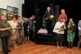 Na scenie stroi mężczyzna, trzymający w rękach kwiaty. Obok niego można zauważyć dyrektorkę ROK-u. Za nimi znajdują się głośniki i czarna kotara. Przed sceną znajdują się kobiety, trzymające w rękach kwiaty. Na ścianie powieszone są obrazy.