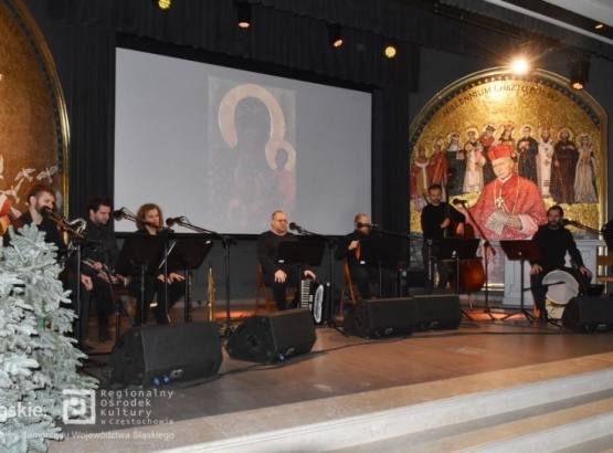 Zespół muzyczny na scenie śpiewający kolędy. Na ścianach sceny sztuka religijna a na ekranie projektora obraz Matki Boskiej.