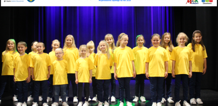 Grupa dzieci w żółtych koszulkach występujących na scenie. Na górze logo sponsorów.