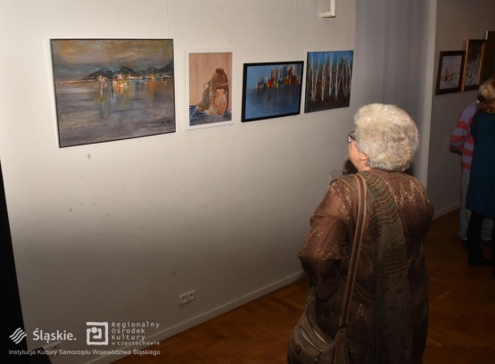 Seniorka z zaciekawieniem przygląda się obrazom wywieszonym na ścianie. W tle widać kawałek innej osoby - kolejnego uczestnika wystawy.