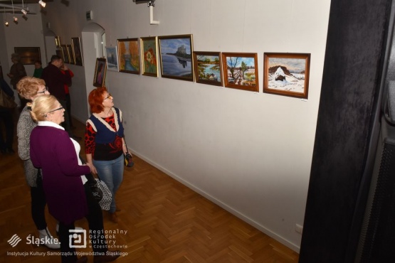 Trójka seniorek ustawionych bokiem do zdjęcia przygląda się obrazom na ścianie. W tle widać inną grupkę uczestników wystawy.