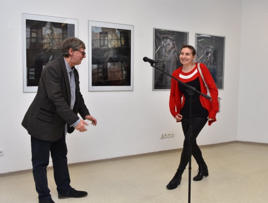 Wystawa fotografii Sławomira Jodłowskiego - organizatorzy przemawiają przy mikrofonie do uczestników . W tle widoczne zdjęcia zawieszone na ścianie.
