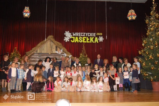 Grupowe zdjęcie dzieci biorących udziały w Jasełkach wraz z opiekunami i organizatorami. Niektórzy z nich prezentują otrzymane dyplomy. W tle stajenka betlejemska oraz choinka.