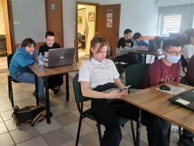 Uczestnicy zajęć z grafiki komputerowej siedzą na sali przy stołach na których stoją laptopy.