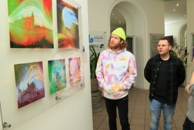 Mężczyźni oglądający obrazy. Na ścianach wiszą obrazy. Jeden z mężczyzn jest ubrany w kolorową bluzę i jasnozieloną czapkę.