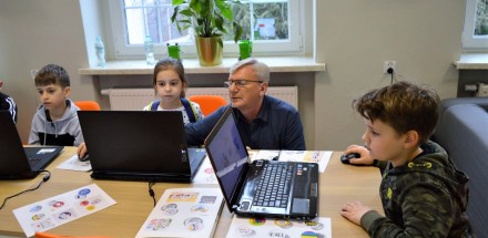 Prowadzący zajęcia instruuje dzieci siedzące przy laptopach. 