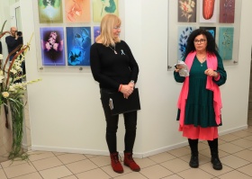 Dwie kobiety stoją obok siebie. W tle wiszą kolorowe obrazy. Jedna z kobiet trzyma w dłoni sreberko.