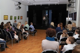 Zdjęcie panorama sali w której odbywa się konkurs- widoczna publiczność oraz uczestniczka stojąca na scenie, która recytuje poezję.