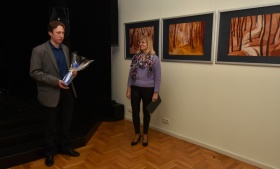 Kobieta stoi obok wystawy z obrazami. W lewej ręce trzyma teczkę. Z lewej strony widoczna mała cześć balonu.