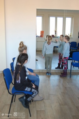 Dzieci ćwiczą aktorstwo przed lustrem, pozostałe uczestniczki siedzą na krzesłach i przyglądają się.