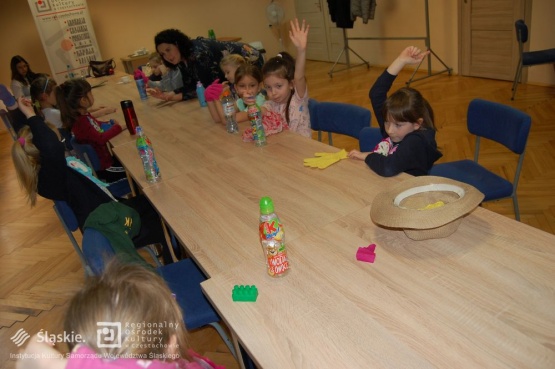 Dzieci siedzą przy stole, niektóre z nich mają podniesione ręce chcąc zadać pytanie prowadzącej warsztaty.