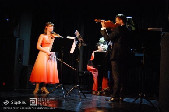 Wieczór Muzyki Kameralnej - koncert skrzypiec (widoczna kobieta i mężczyzna ze skrzypcami) w tle kobieta gra na pianinie.