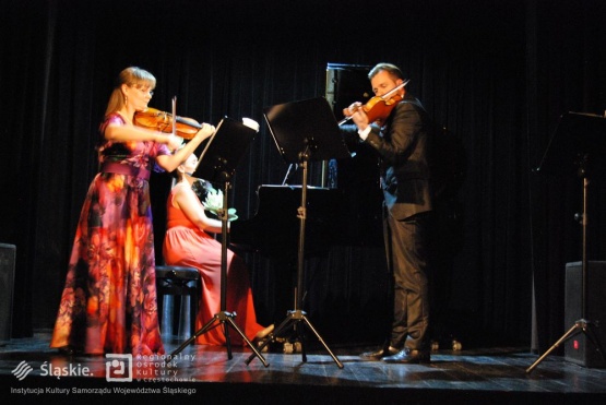 Na scenie występuje kobieta i mężczyzna, którzy grają na skrzypcach, w tle kobieta grająca na pianinie.
