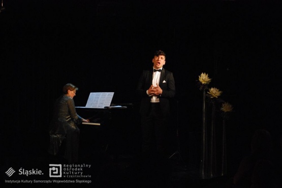 .Męzczyzna z złożonymi rękami śpiewa na scenie ubrany w czarny smoking. Obok niego kobieta grająca na pianinie. Po prawej są widoczne białe kwiaty.