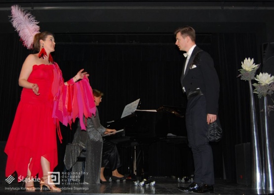 Para występuje na scenie. Kobieta jest ubrana w czerwoną suknię z jasno różową opaską na głowie, a mężczyzna w czarny frak. W tle gra kobieta na pianinie