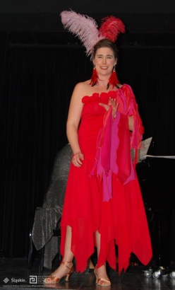 Kobieta na scenie w czerwonej sukni w opasce z piór na głowie wykonuje utwór. Na twarzy można zauważyć mocne zaangażowanie i lekki uśmiech. Ma uniesioną dłoń przed siebie na wysokości klatki piersiowej.