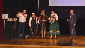 Mężczyzna w stroju ludowym śpiewa do mikrofonu, obok na harmonii gra kobieta w stroju ludowym, z drugiej strony sceny mężczyzna grający na flecie.