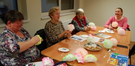 Uczestniczki zajęć plastycznych siedzą przy drewnianym stole i wykonują kolorowe kwiaty z papieru.