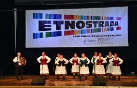 Kobiety w strojach ludowych tańczą i śpiewają na scenie, obok na akordeonie gra mężczyzna ubrany w strój ludowy.