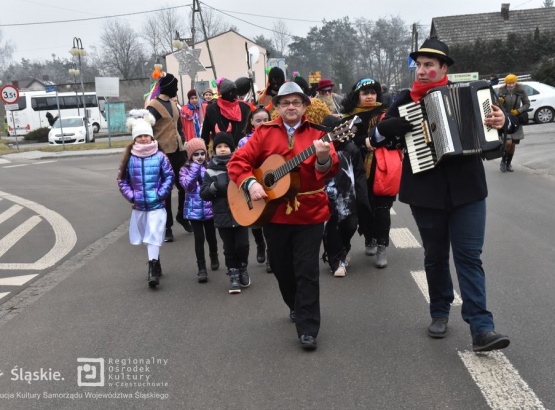Uczestnicy wydarzenia idą w orszaku po ulicy - na czele mężczyzna z akordeonem oraz mężczyzna z gitarą, za nimi idą dzieci.
