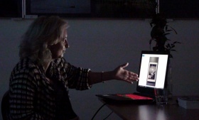 Katarzyna Łata prezentująca swój wykład. Ręką wskazuje na ekran monitora. Zdjęcie zrobione przy zgaszonych światłach.