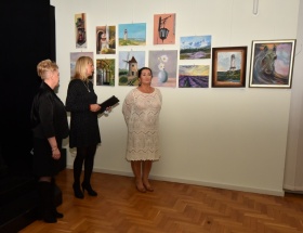Trzy kobiety, które prowadzą wystawę stoją obok siebie na tle powieszonych obrazów. Kobiety są ubrane w suknie, jedna z nich trzyma teczkę.