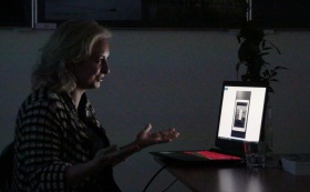Katarzyna Łata prowadzi swoje spotkanie. Na twarzy maluje się radość, gestykuluje przed monitorem. Siedzi na krześle. Zdjęcie zostało zrobione przy zgaszonych światłach.