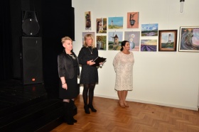 Trzy kobiety prowadzące wystawę ubrane w eleganckie sukienki mają skierowany wzrok na publiczność. Stoją przed ścianą z obrazami. Widoczny obok głośnik.