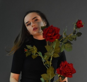 Kobieta ubrana na czarno ustawiona przodem do aparatu, w rękach trzyma czerwone róże.