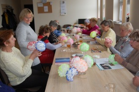Uczestnicy warsztatów plastycznych siedzą przy stole i tworzą prace - papierowe kwiaty w różnych kolorach.