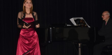 Kobieta stoi na scenie obok fortepianiu.