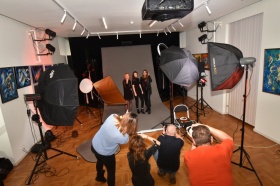 Sesja zdjęciowa w studio - na pierwszym planie fotografowie wykonują zdjęcia trzem kobietom. W tle widoczny sprzęt do oświetlenia i parasole.