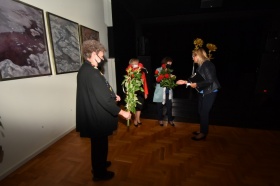 Kobieta wręcza kwiaty innym uczestnikom wystawy. W tle czarna kotara, po lewej stronie wiszą na ścianach dwa obrazy,